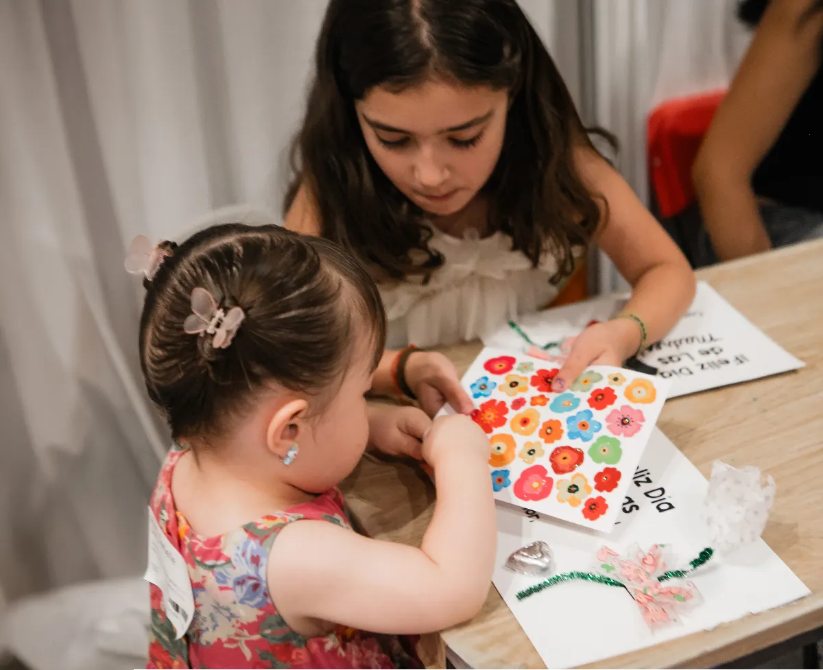 Dos niñas decoran juntas una tarjeta con calcomanías de flores durante Mosaic Kids