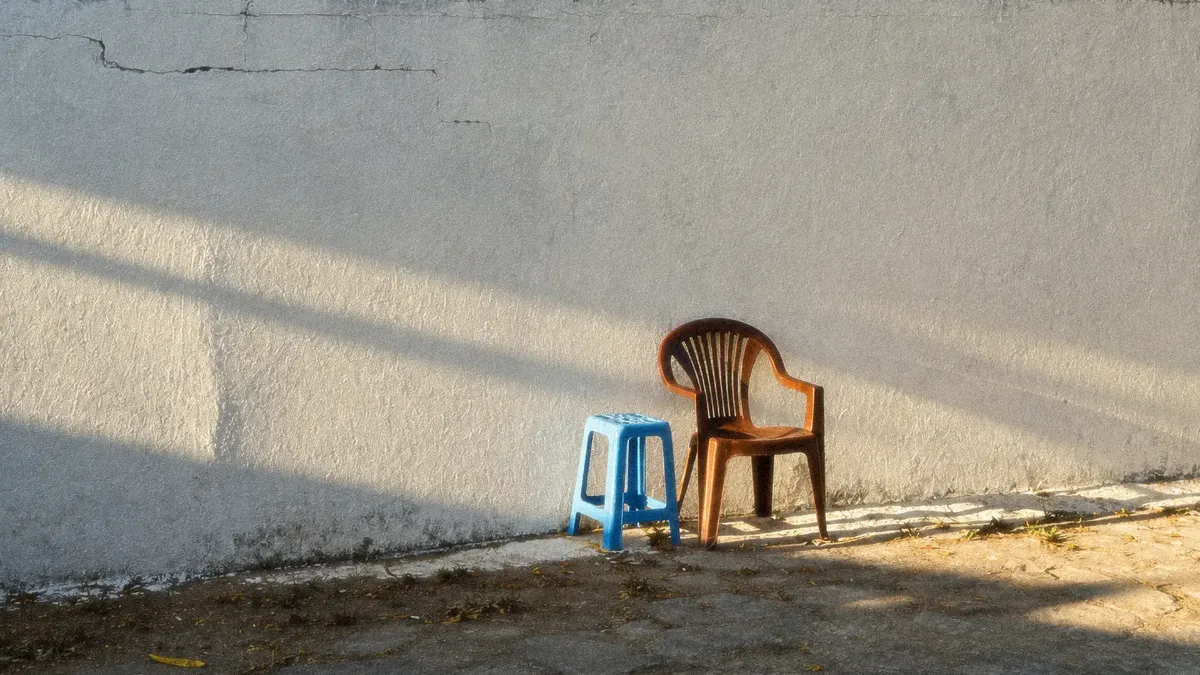 Una silla café y un banco azul de plástico, juntos frente a un muro blanco con luz de tarde
